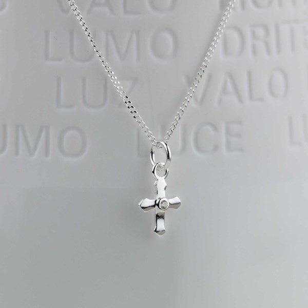 A silver cross pendant necklace with a small clear gemstone in the center, hanging from a delicate silver chain against a light gray background with embossed Spanish text.