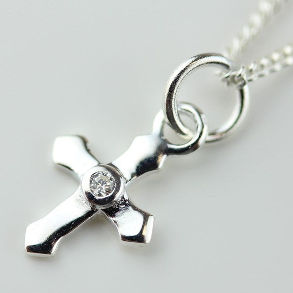A polished silver cross pendant necklace with a small clear gemstone set in the center, attached to a silver chain on a light gray background.