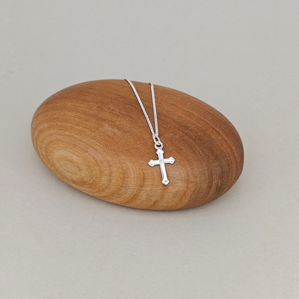 A silver cross pendant necklace on a delicate silver chain, displayed on a smooth, polished wooden stone against a neutral light gray background.