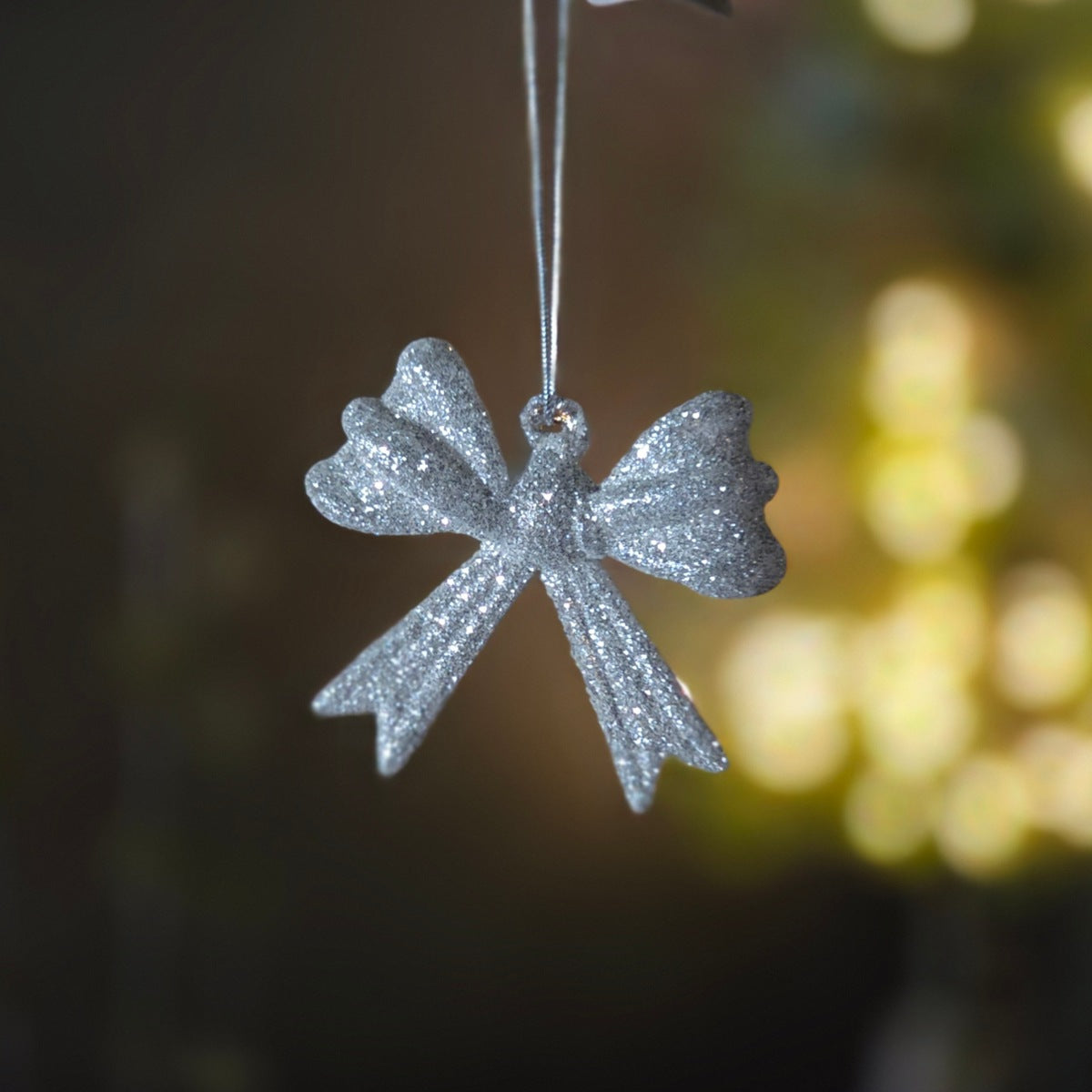A silver glitter Christmas ornament shaped like a bow, hanging from a thin silver string against a dark, softly blurred background with warm yellow bokeh lights.