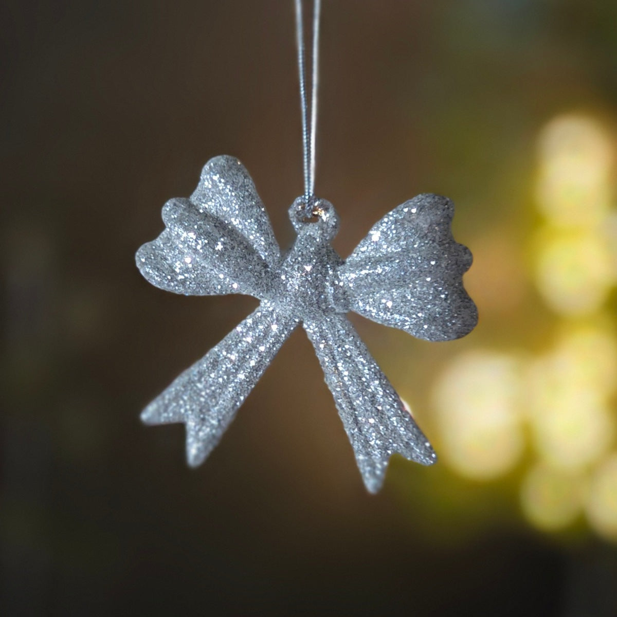 A silver glitter Christmas ornament shaped like a bow, hanging from a thin silver string against a dark, softly blurred background with warm yellow bokeh lights.