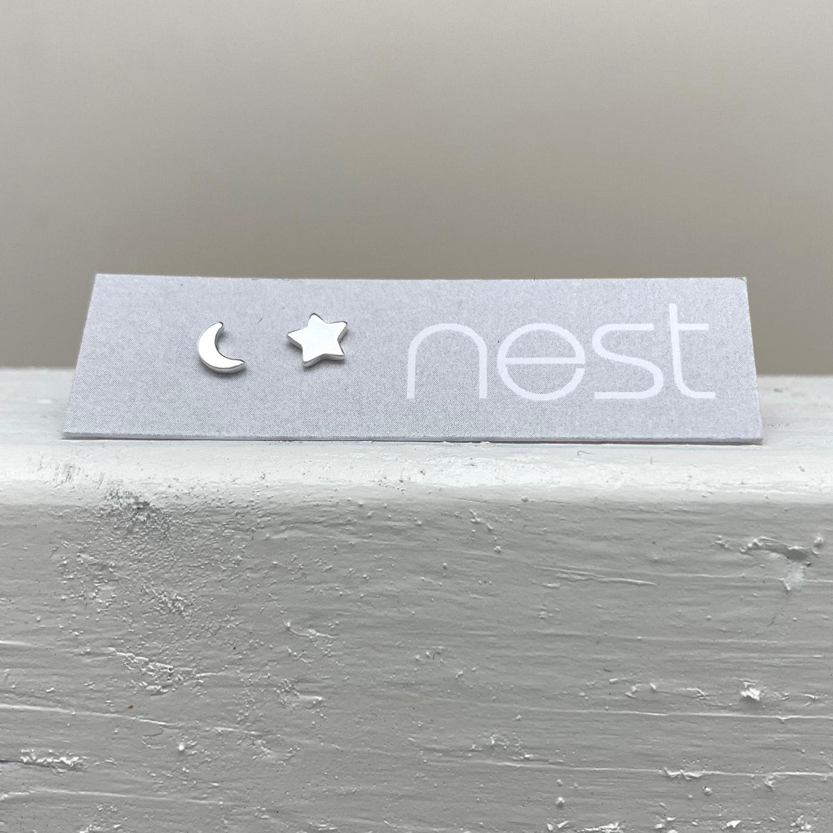 A pair of small silver stud earrings, one crescent moon and one star, displayed on a gray card with the brand name 'nest' printed on it, resting on a textured gray surface.