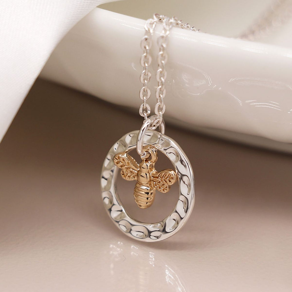 Silver chain necklace with a silver hammered ring and gold bee pendant dangling inside the ring on a beige surface.