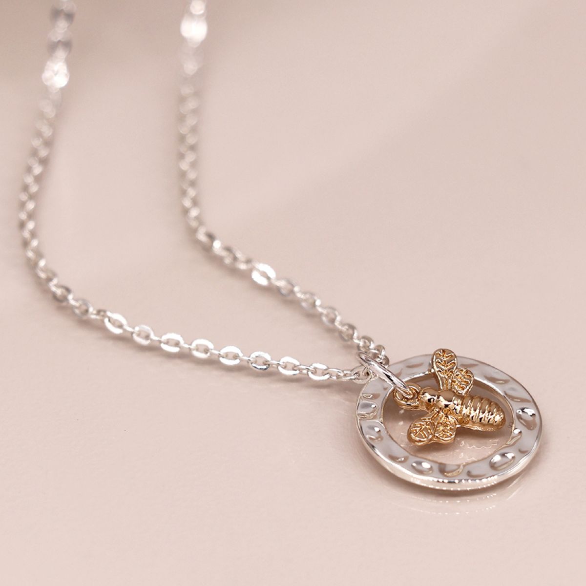 Silver chain necklace with a silver hammered ring and gold bee pendant dangling inside the ring on a beige surface.