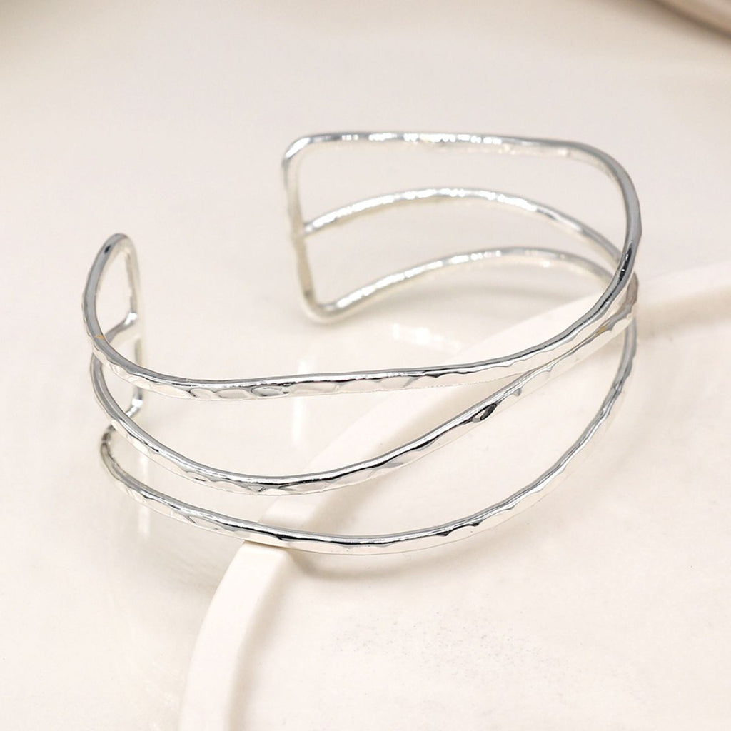 A silver open cuff bangle bracelet with a hammered texture, displayed on a clean, light beige surface.