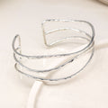 A silver open cuff bangle bracelet with a hammered texture, displayed on a clean, light beige surface.