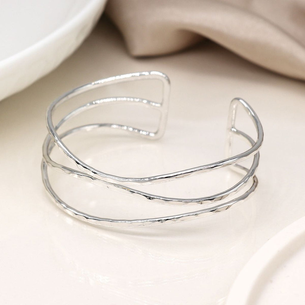 A silver open cuff bangle with a hammered texture, displayed on a light cream surface with soft-focus white and beige background elements.
