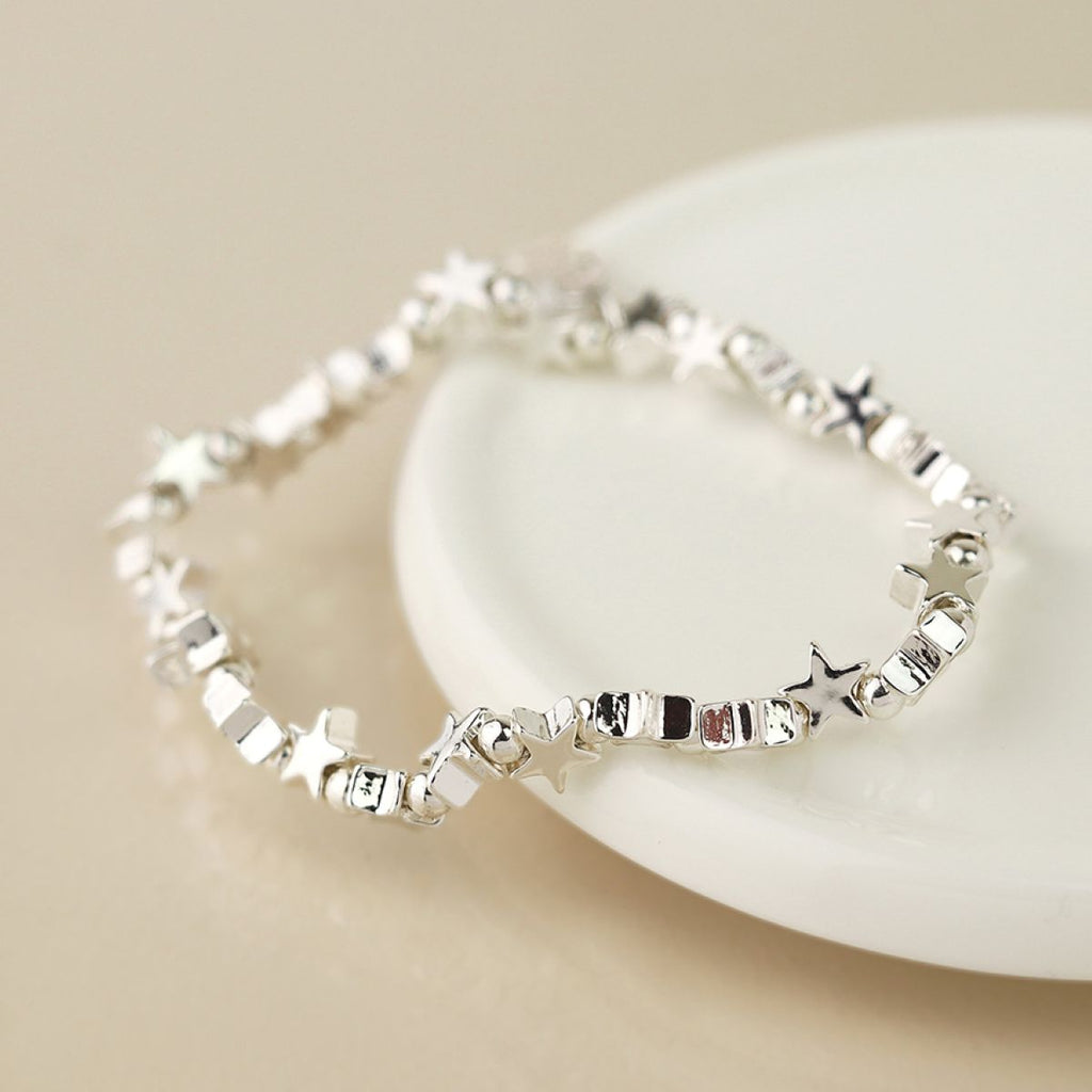 A silver bracelet with connecting star and bead charms, resting on a white ceramic dish against a warm beige background.