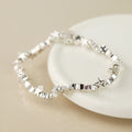 A silver bracelet with connecting star and bead charms, resting on a white ceramic dish against a warm beige background.
