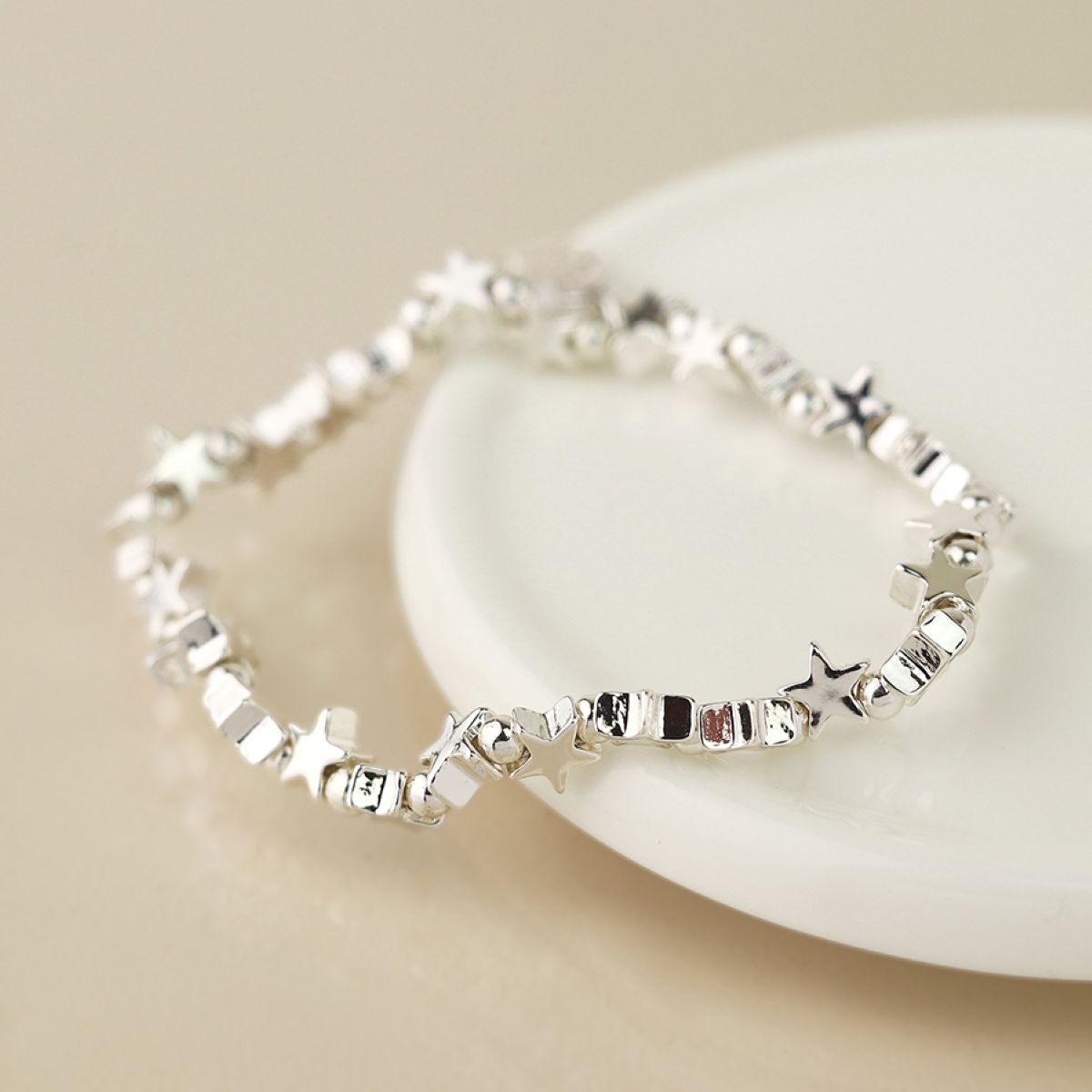 A silver bracelet with connecting star and bead charms, resting on a white ceramic dish against a warm beige background.