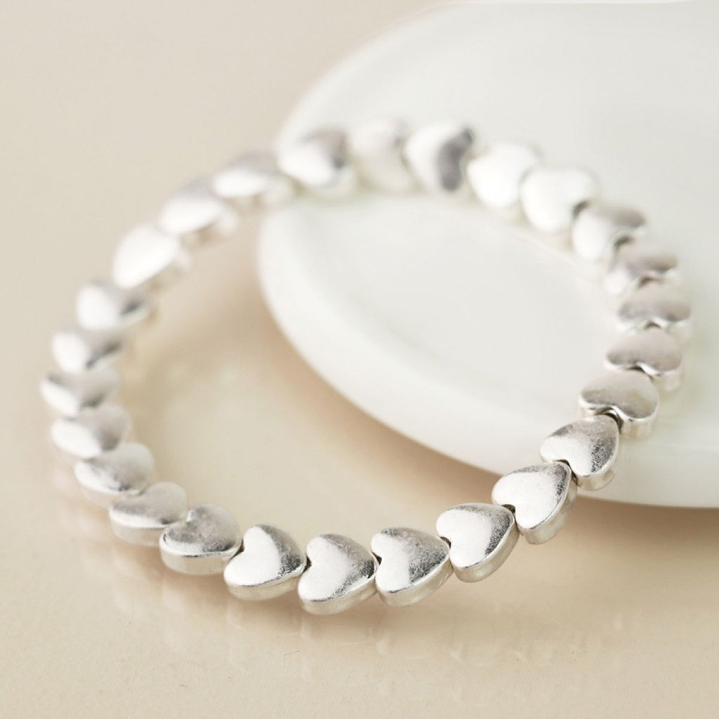 A silver heart-link stretch bracelet resting on a white ceramic dish against a warm beige background.