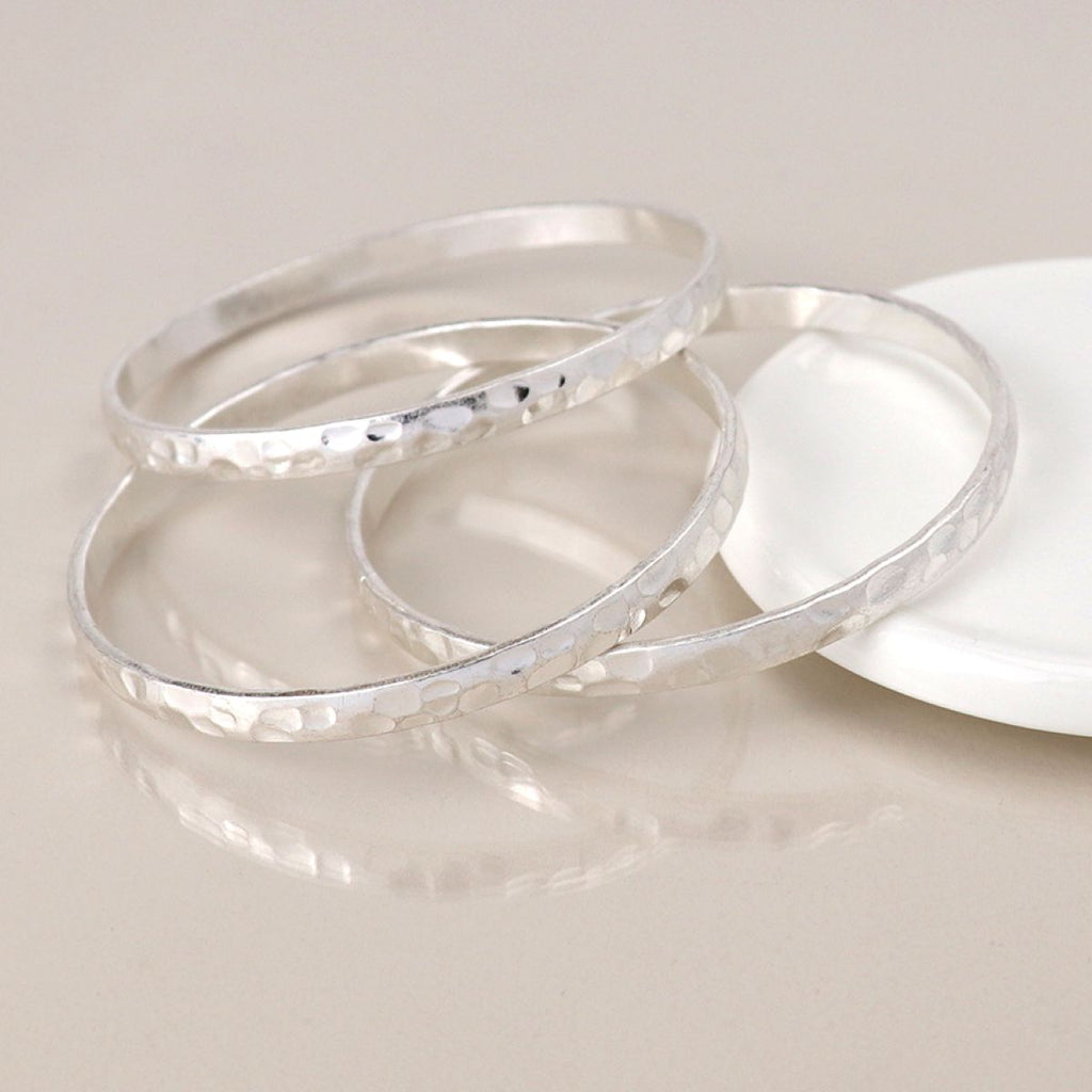 Three hammered silver bangles stacked together on a light beige reflective surface, with a white round dish partially visible in the background.