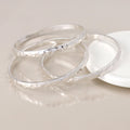 Three hammered silver bangles stacked together on a light beige reflective surface, with a white round dish partially visible in the background.