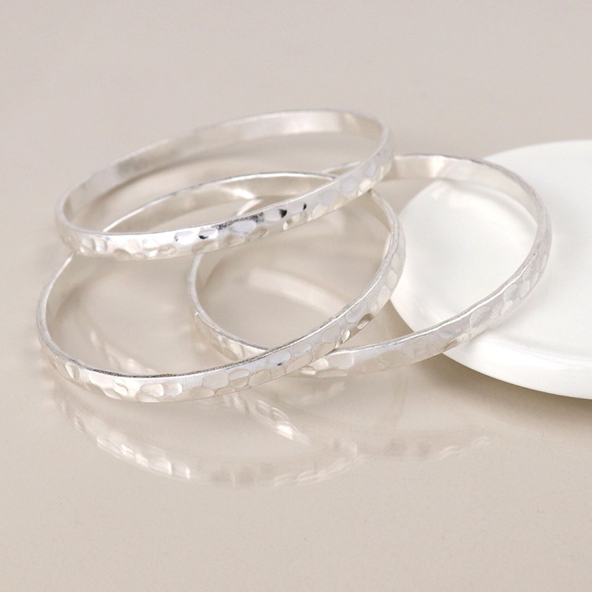 Three hammered silver bangles stacked together on a light beige reflective surface, with a white round dish partially visible in the background.