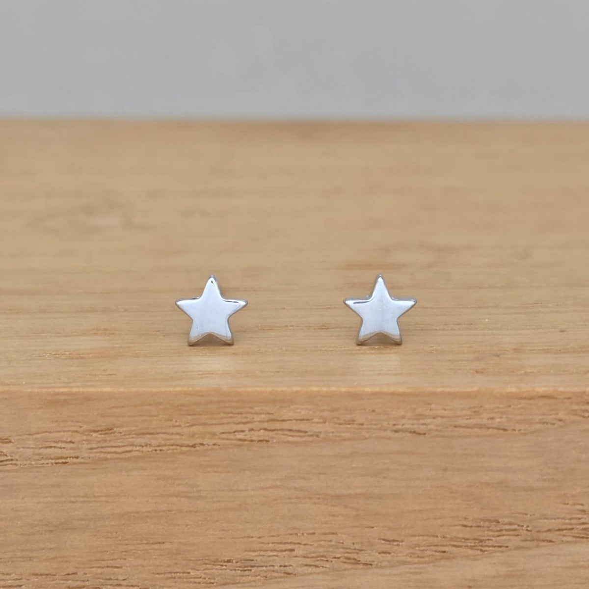 A pair of small silver star stud earrings resting on a light brown wooden surface against a neutral gray background.