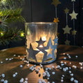 Decorative glass candle holder with star metal cutouts on a wooden surface with a Christmas tree and stars in the background.