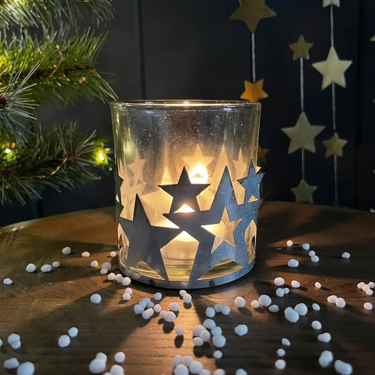 Decorative glass candle holder with star metal cutouts on a wooden surface with a Christmas tree and stars in the background.