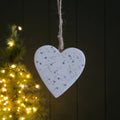 White heart-shaped ornament with little silver stars and dots hanging against a dark wooden background with a Christmas tree in the foreground.