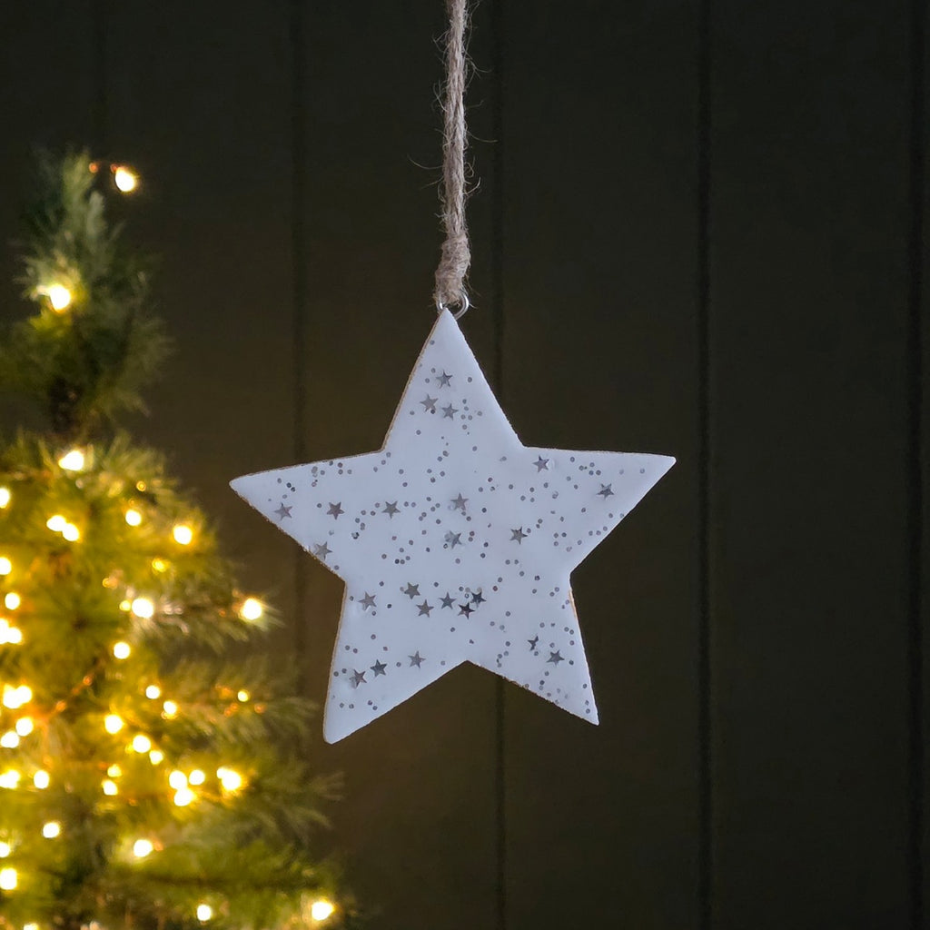 White star-shaped ornament with little silver stars and dots hanging against a dark wooden background with a Christmas tree in the foreground.
