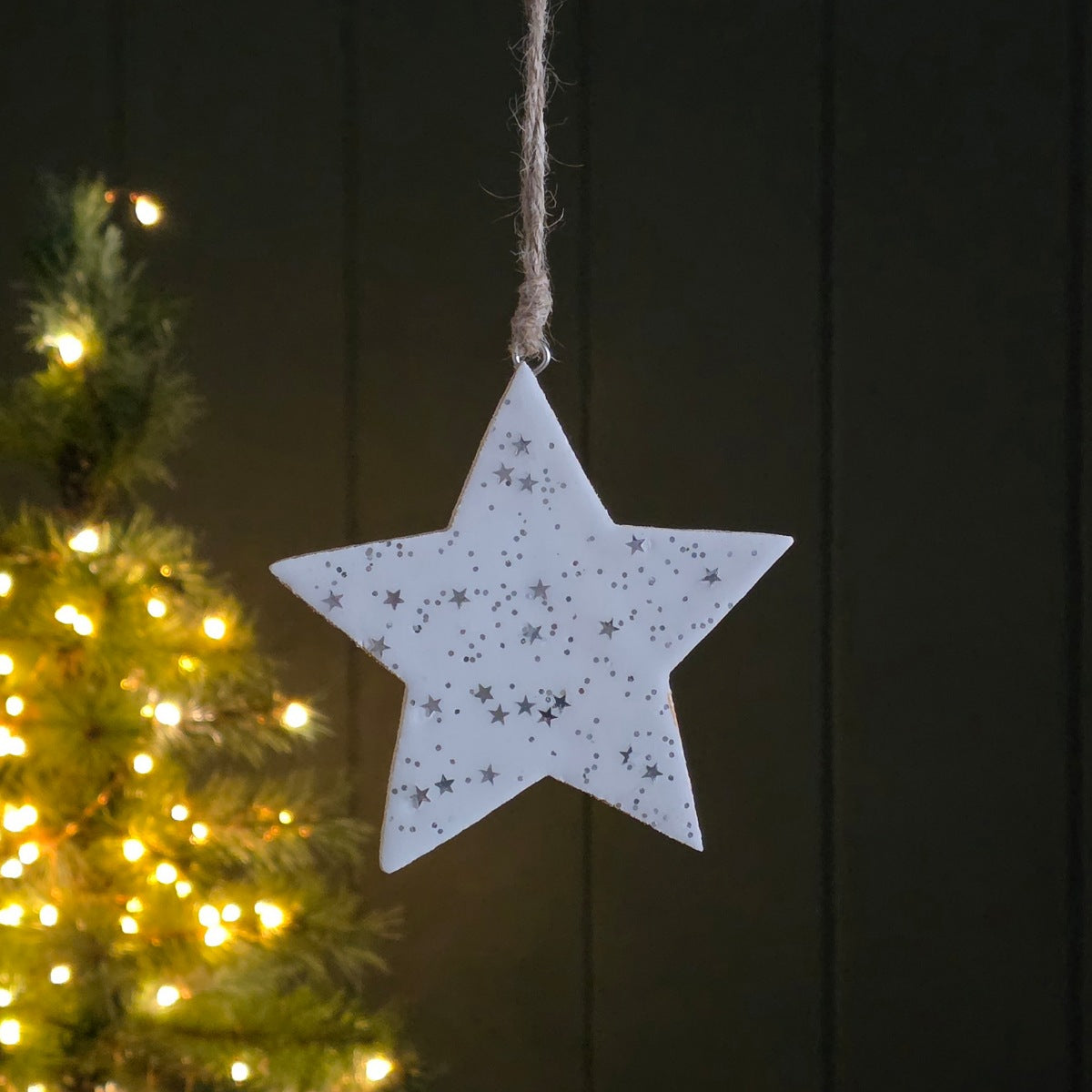 White star-shaped ornament with little silver stars and dots hanging against a dark wooden background with a Christmas tree in the foreground.