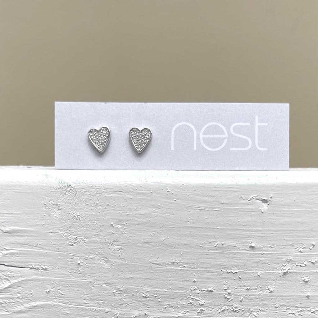 A pair of small silver heart stud earrings displayed on a white card printed with the brand name 'nest', set against a neutral beige background and a textured white surface.