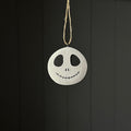A circular white hanging ornament with a black hand-drawn face resembling a skull and stitched smile, suspended by a natural twine string against a dark grey panelled background.