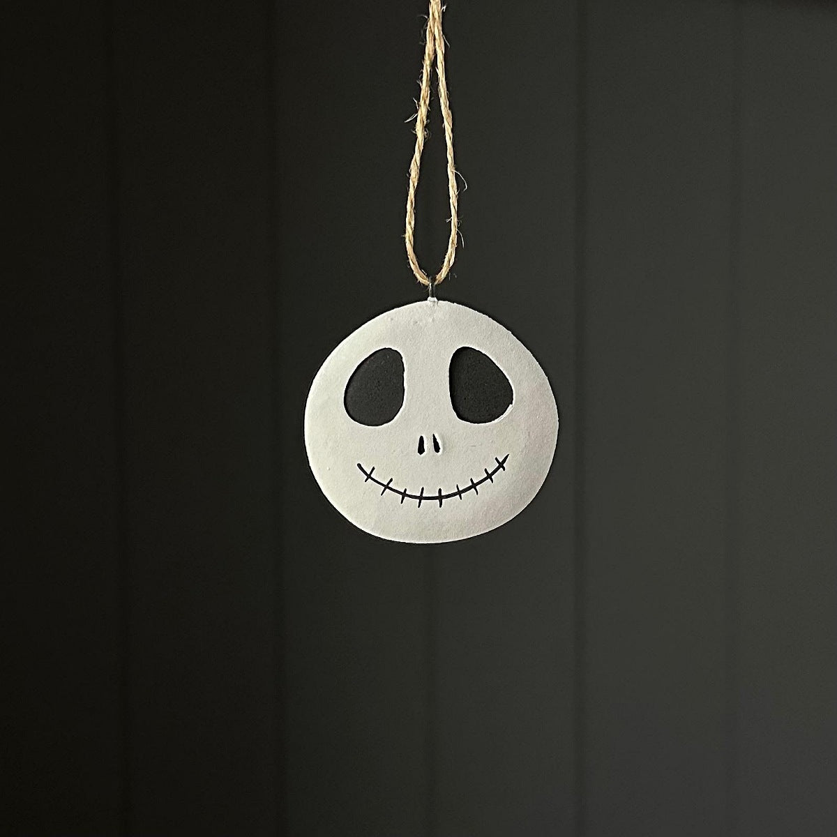 A circular white hanging ornament with a black hand-drawn face resembling a skull and stitched smile, suspended by a natural twine string against a dark grey panelled background.