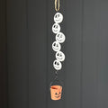 A metal hanging Halloween decoration made of five stacked white skeleton faces with black eyes and mouths, suspended above a small orange bucket with a black handle, against a dark grey panelled background.