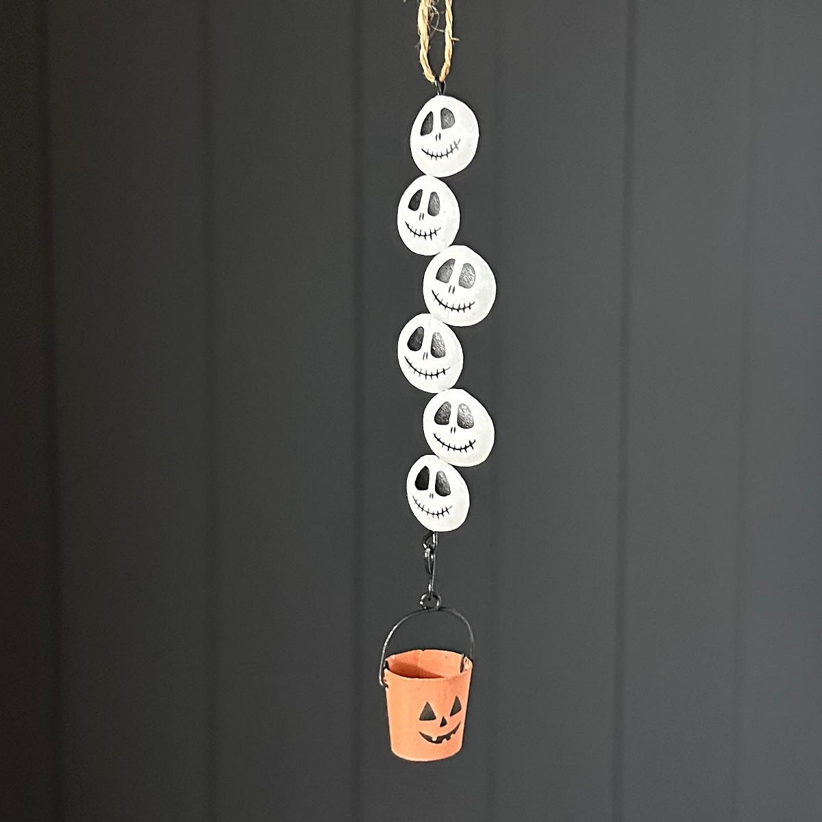 A metal hanging Halloween decoration made of five stacked white skeleton faces with black eyes and mouths, suspended above a small orange bucket with a black handle, against a dark grey panelled background.