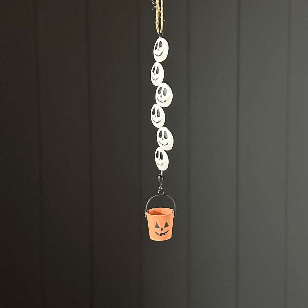 A Halloween hanging decoration featuring a vertical string of white circular plates printed with black jack-o'-lantern faces, ending in a small orange bucket with a carved face.