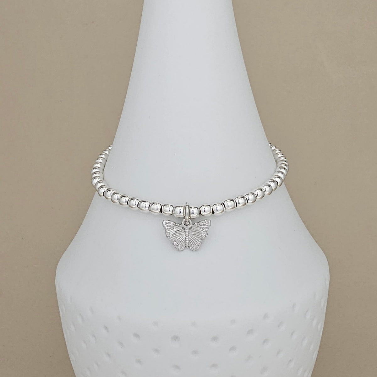 A silver beaded bracelet with a small butterfly charm, displayed on a white cone-shaped jewelry stand against a beige background.