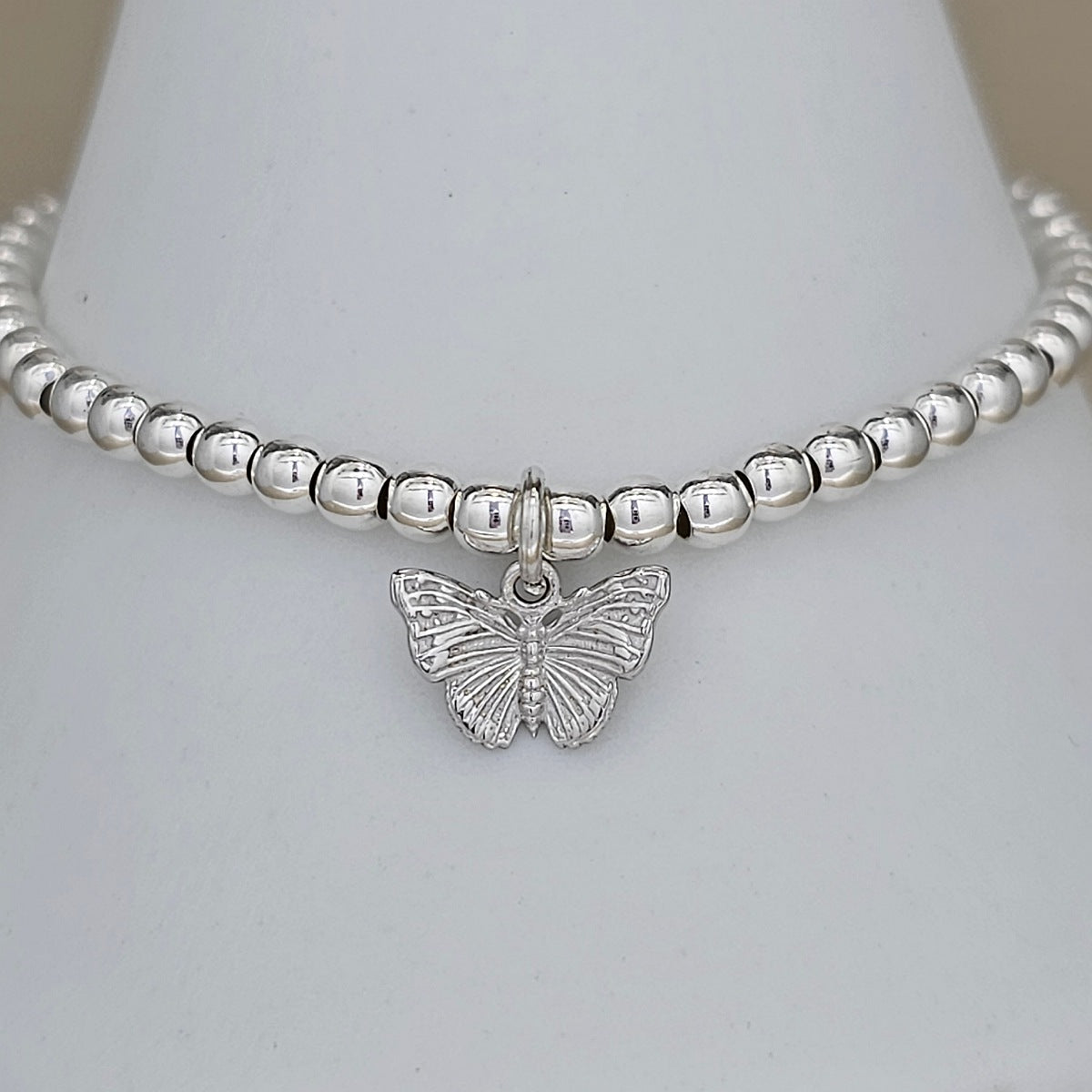 A silver beaded bracelet with a small butterfly charm, displayed on a white cone stand.