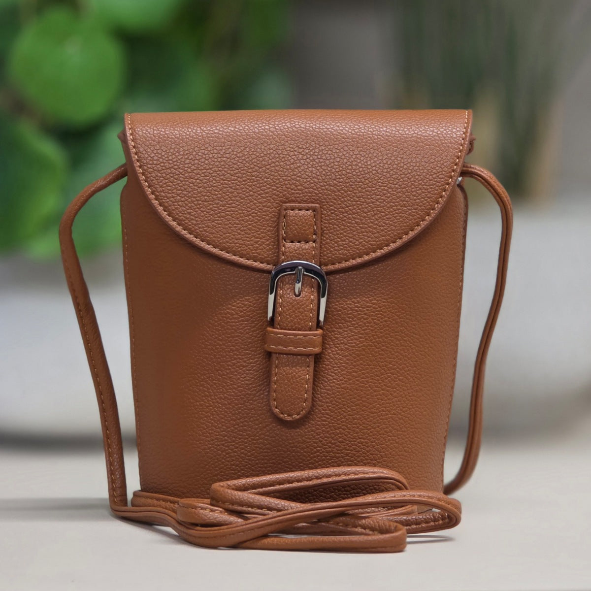 Brown leather crossbody bag with a buckle on a light surface with a blurred green background