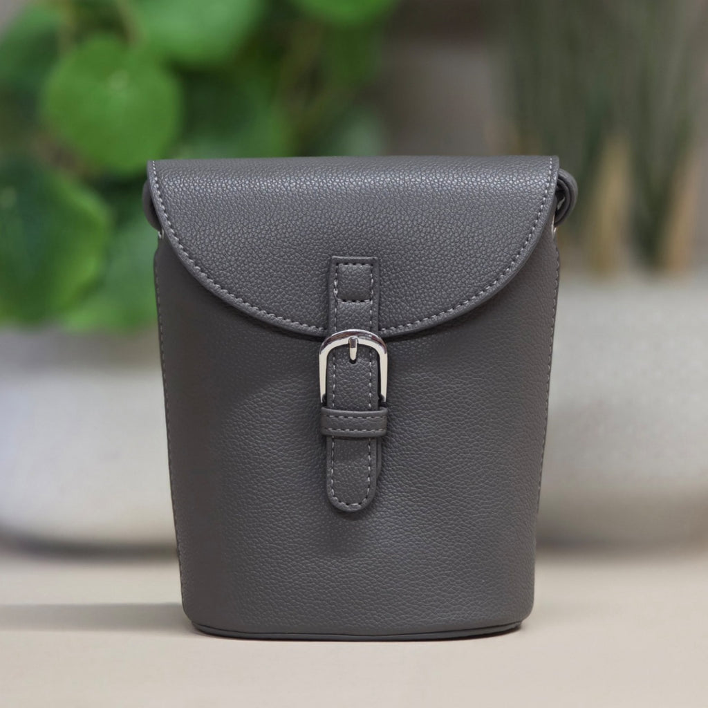 A dark gray pebbled leather phone pouch with a front flap and silver buckle, set against a softly blurred background of green plants and a light surface.