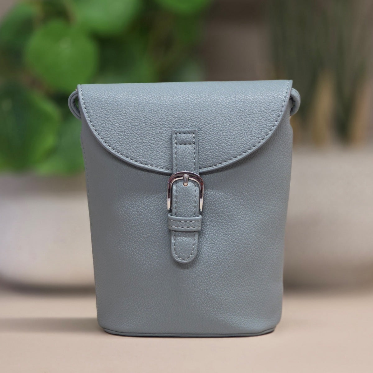 A light blue pebbled leather crossbody bag with a front flap and silver-tone buckle, photographed against a softly blurred indoor background with greenery.