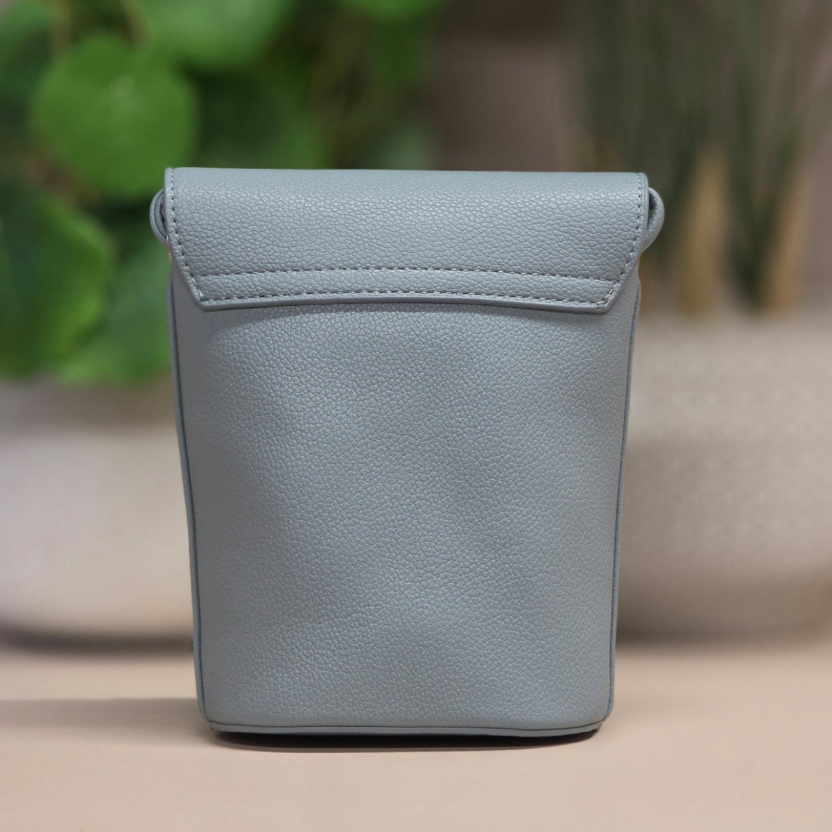 A light blue pebbled leather pouch with a top flap and stitched edge, standing upright on a beige surface with a blurred green plant in the background.