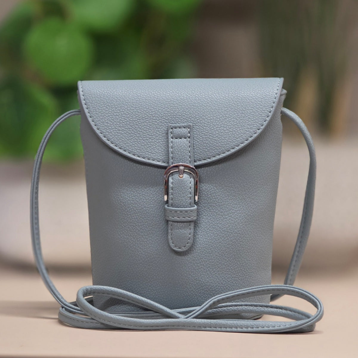 A light blue pebbled leather crossbody phone bag with a flap closure and silver buckle, shown on a neutral surface with a blurred green background.