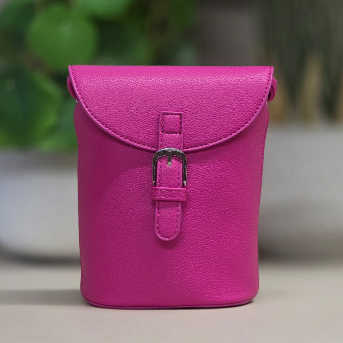A small bright pink pebbled leather pouch with a flap closure and silver buckle, photographed standing upright against a softly blurred indoor background with greenery.