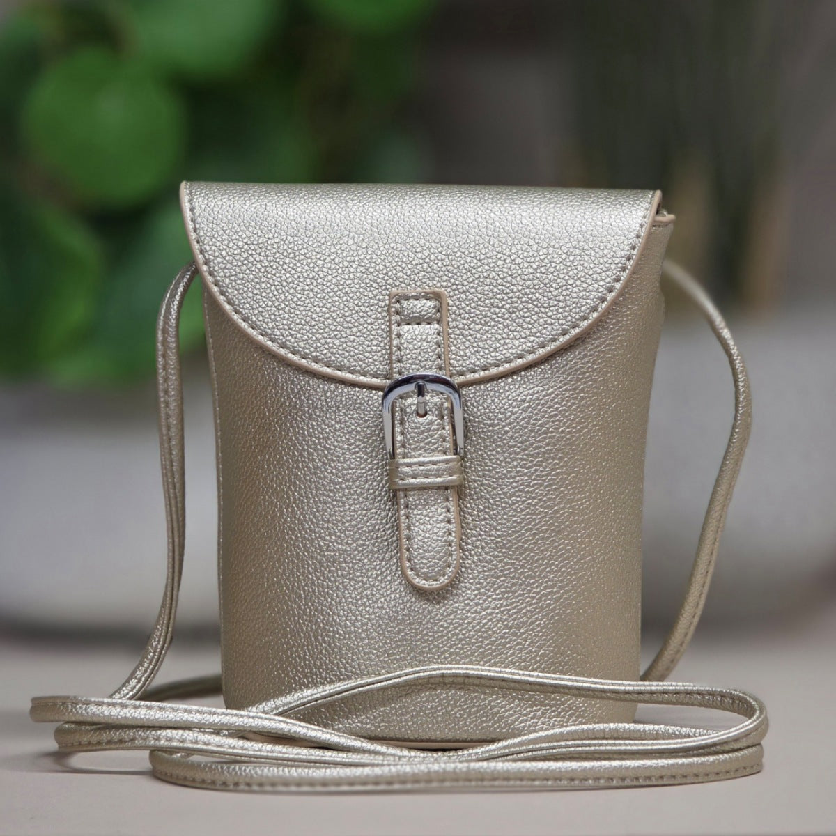 A metallic champagne-gold textured leather crossbody bag with a front flap and buckle closure, resting on a neutral surface with a blurred green background.
