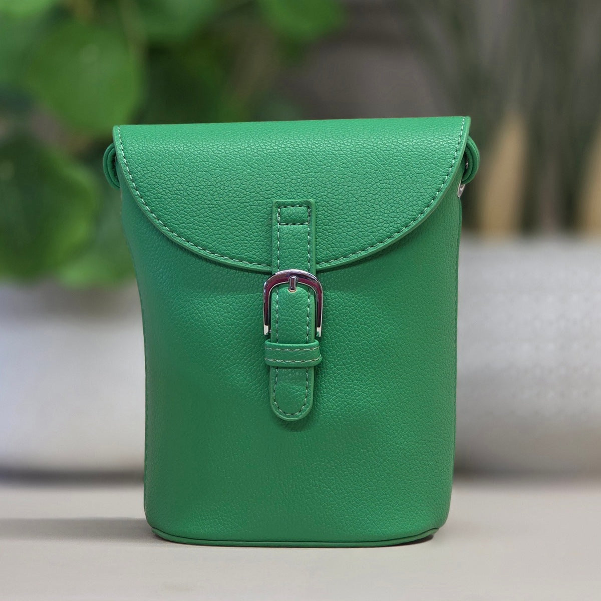 A green pebbled leather crossbody phone pouch with a front flap and silver-tone buckle, shown standing upright against a softly blurred indoor background.