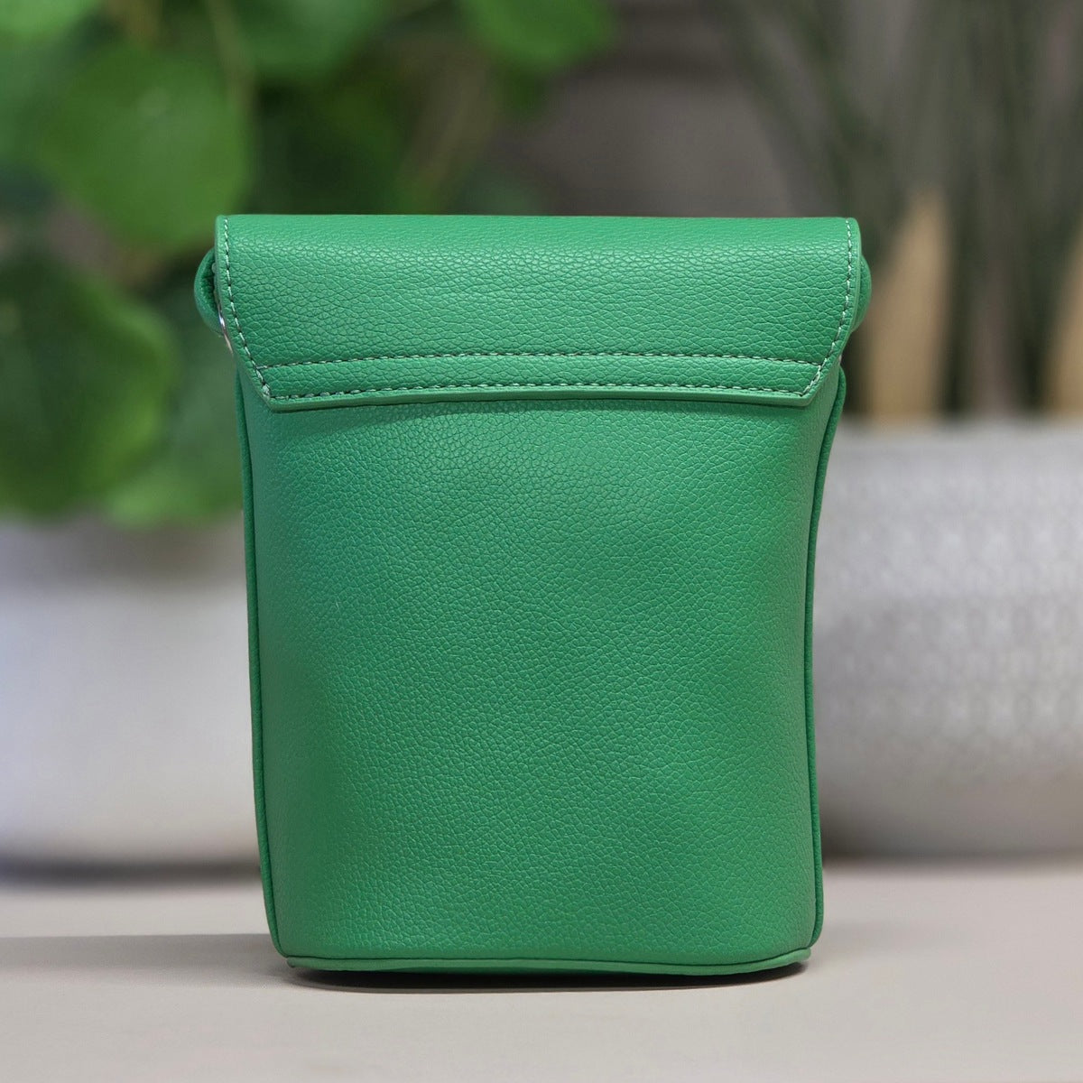A bright green pebbled leather pouch with a top flap and stitched edge, standing upright on a light surface with a blurred indoor background.