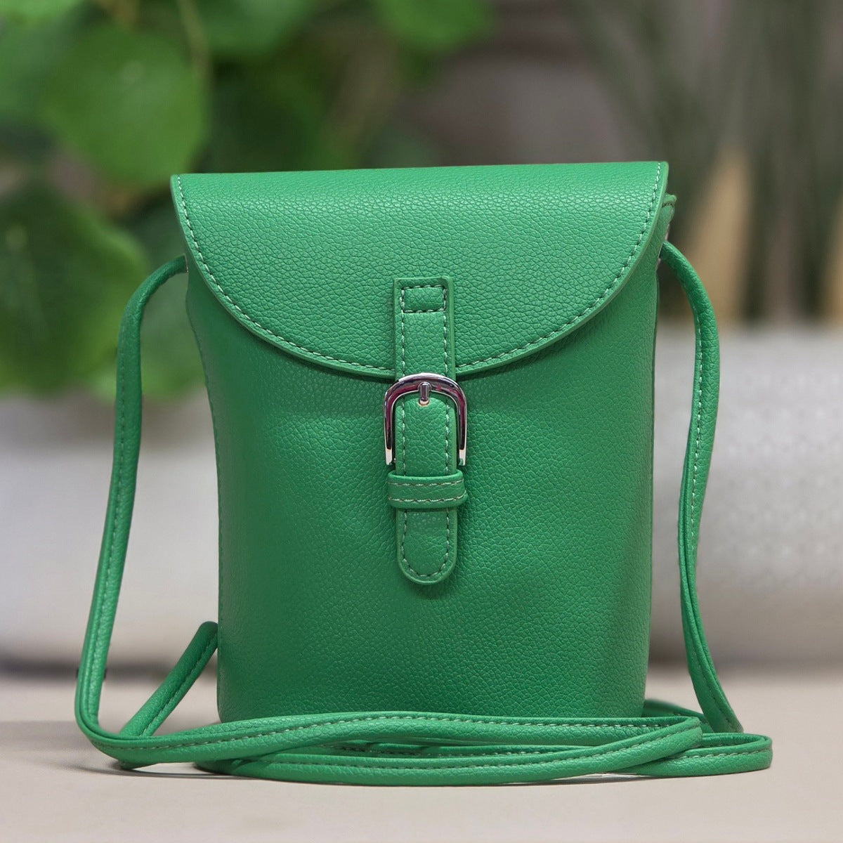 A bright green pebbled leather crossbody bag with a flap front, silver buckle closure, and thin shoulder strap, photographed against a softly blurred indoor background with greenery.