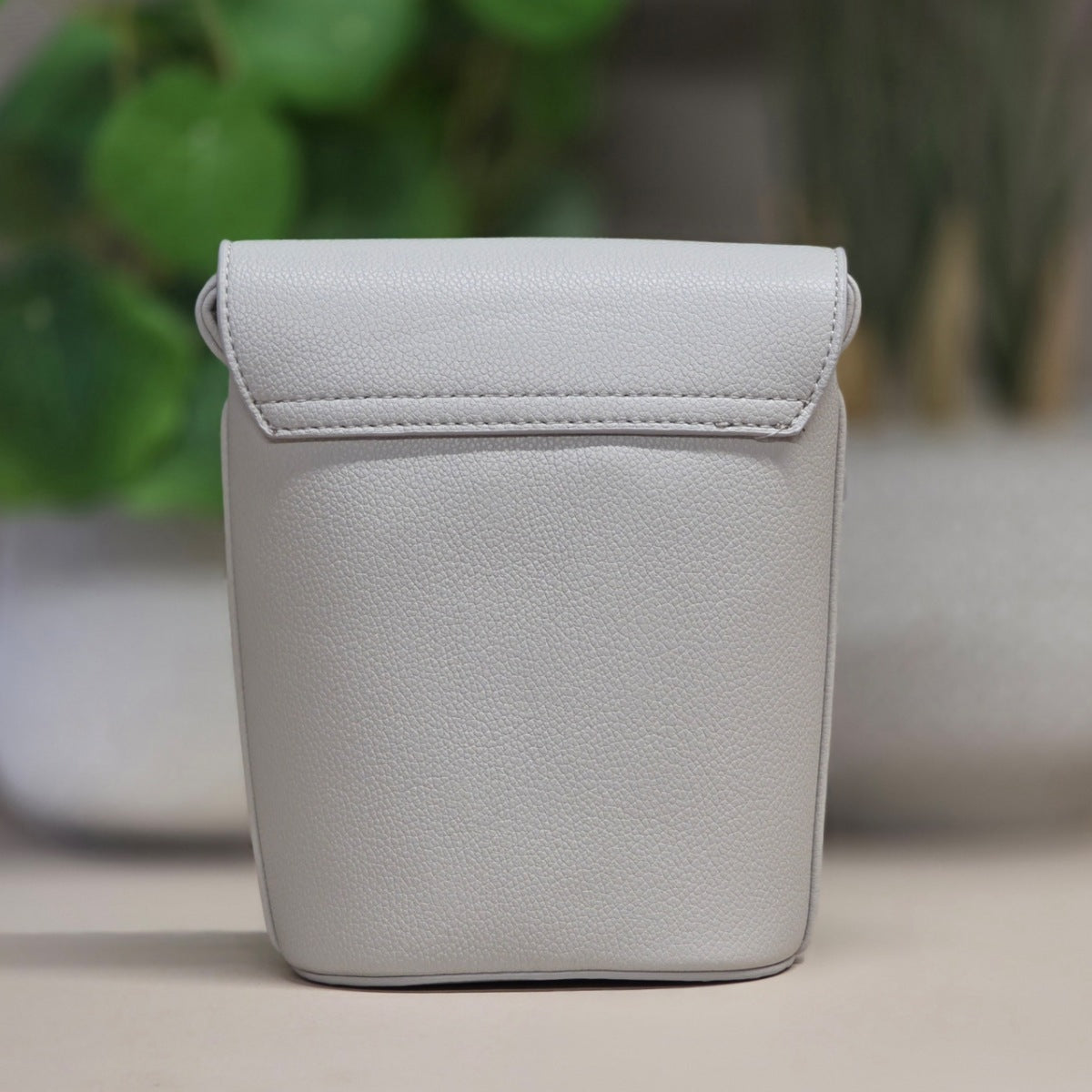 A light gray pebbled leather pouch with a top flap and stitched edge, standing upright on a neutral surface with a blurred green plant in the background.