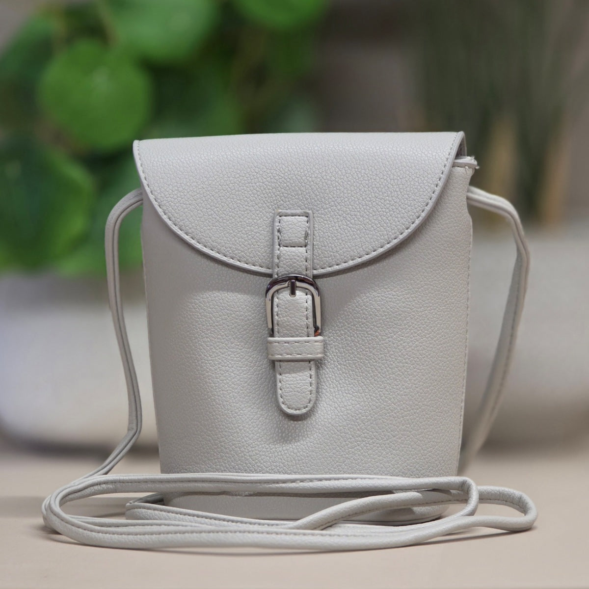 A light gray pebbled faux-leather crossbody bag with a front flap, buckle closure, and thin shoulder strap, shown against a softly blurred indoor background with green plants.
