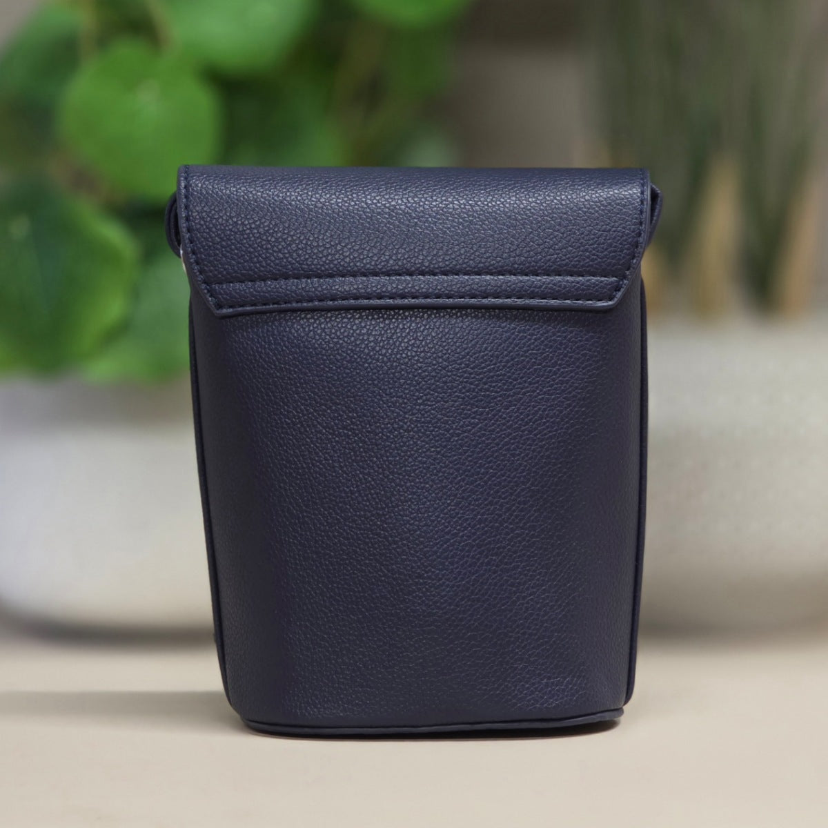 A navy blue textured leather pouch with a flap closure, standing upright on a light beige surface with a blurred green plant in the background.