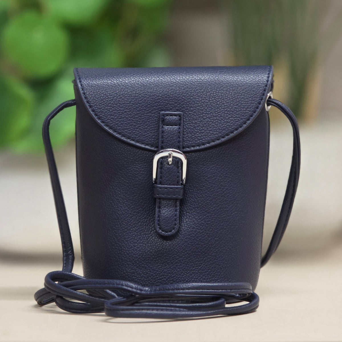 A navy blue pebbled leather crossbody bag with a flap closure and silver buckle, shown on a neutral surface with a blurred green background.