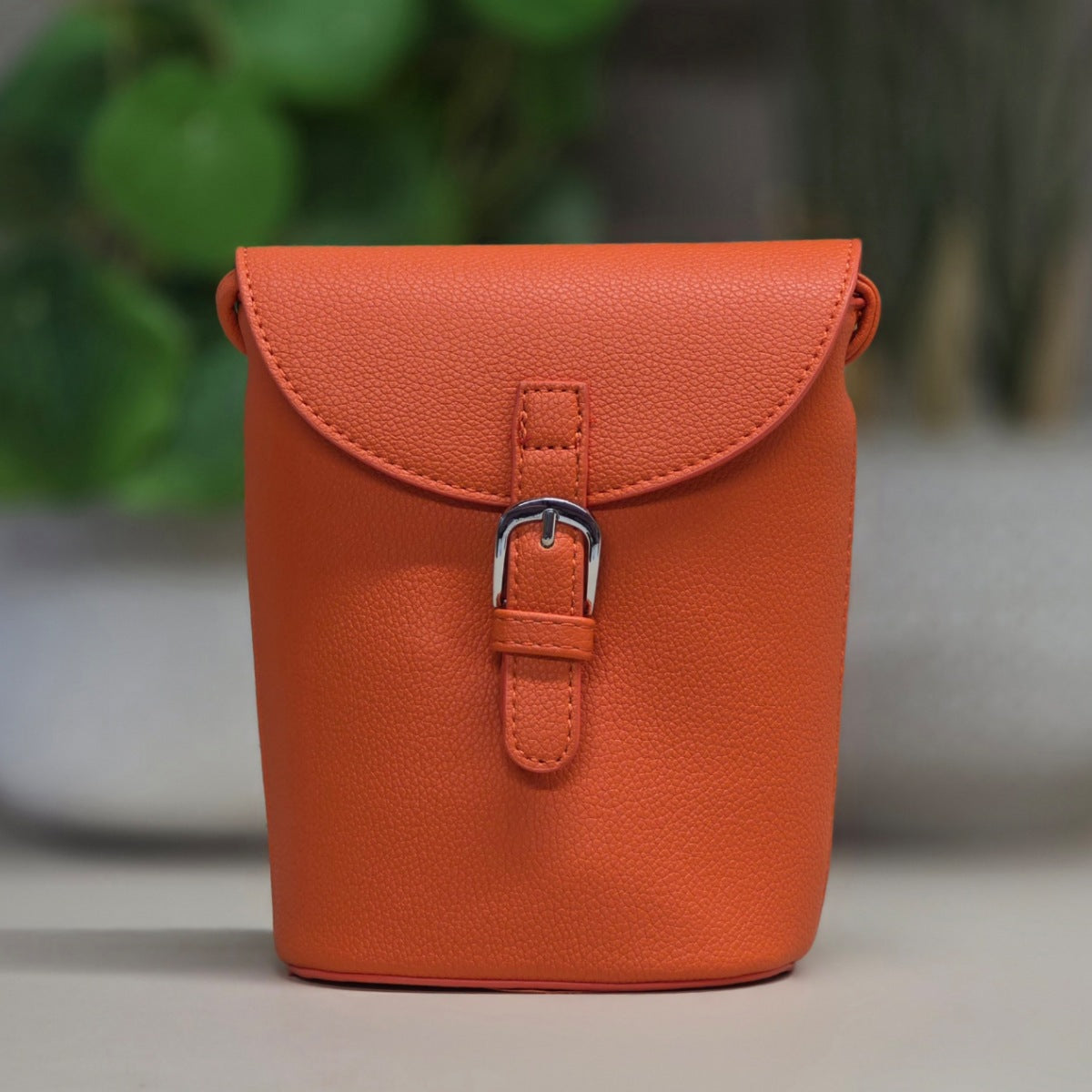 An orange textured leather pouch bag with a flap closure and silver buckle, set against a blurred background of green plants and a gray surface.