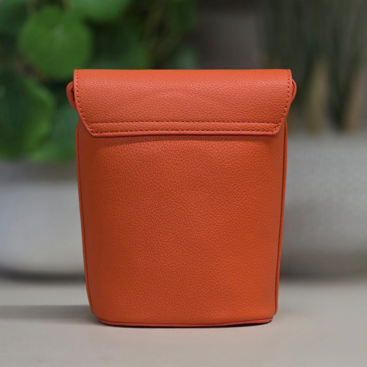 An orange textured leather pouch with a fold-over flap, standing upright on a light surface with a blurred green and gray background.