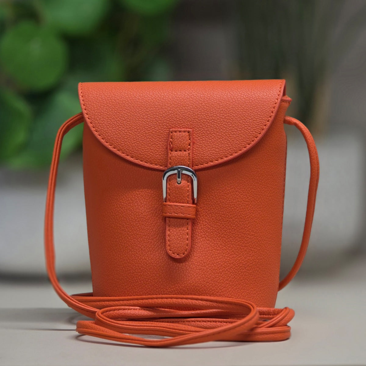 An orange pebbled leather crossbody bag with a flap closure and silver buckle, shown on a neutral surface with a blurred green background.