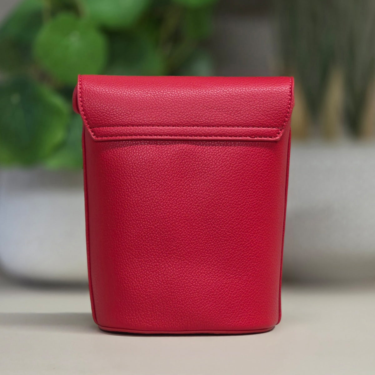 A bright red textured leather pouch with a flap closure, standing upright on a light surface with a blurred green plant in the background.